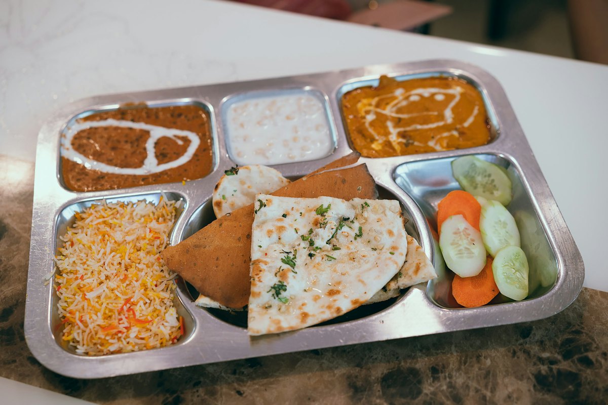 A steel compartment tray with rice, dal, curry, paratha and cucumber and carrot sticks